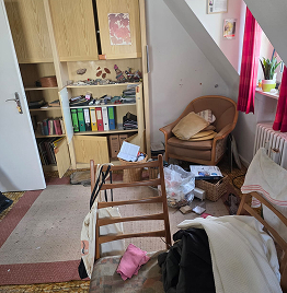 Unaufgeräumtes Zimmer mit umgestürztem Stuhl, verstreuten Gegenständen und überfülltem Regal während einer Entrümpelung