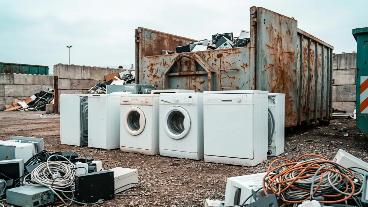 Alte weiße Waschmaschinen und andere Elektrogroßgeräte an einer Sammelstelle für Elektroschrott auf einem Recyclinghof, dahinter ein großer Sammelcontainer
